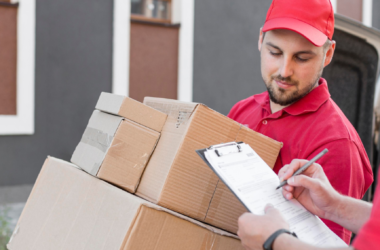 Two delivery workers in red uniforms unload cardboard boxes from a van; one writes on a clipboard nearby.